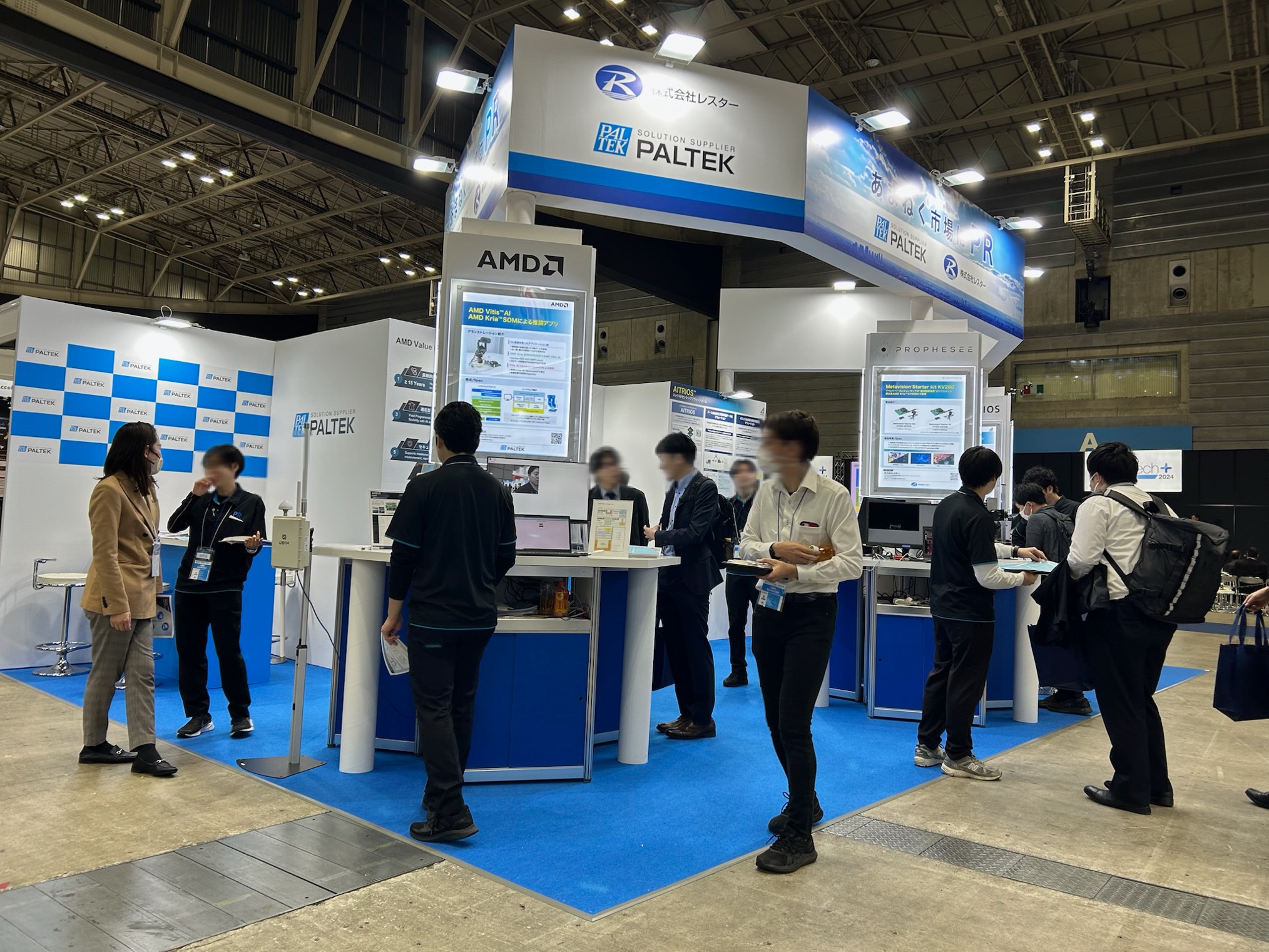 PALTEKとレスターの共催による展示会ブース。白と青を基調としたブースデザインで、各社のロゴが掲示されている。来場者がデモ展示を見学し、スタッフと対話している様子。ブース内には情報パネルやデモンストレーション用のディスプレイが設置されている。