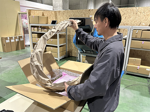 倉庫内で作業員が段ボール箱の上部から紙製緩衝材を持ち上げ、形を整えている様子。背景には保管用の棚とPadPak®Guardian製品が設置されている
